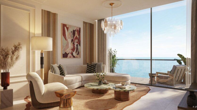 A modern living room in the Château with a curved white sofa, round coffee tables, abstract wall art, and floor-to-ceiling windows overlooking the ocean. Light floods the space, highlighting a chandelier, indoor plants, and neutral-toned décor.