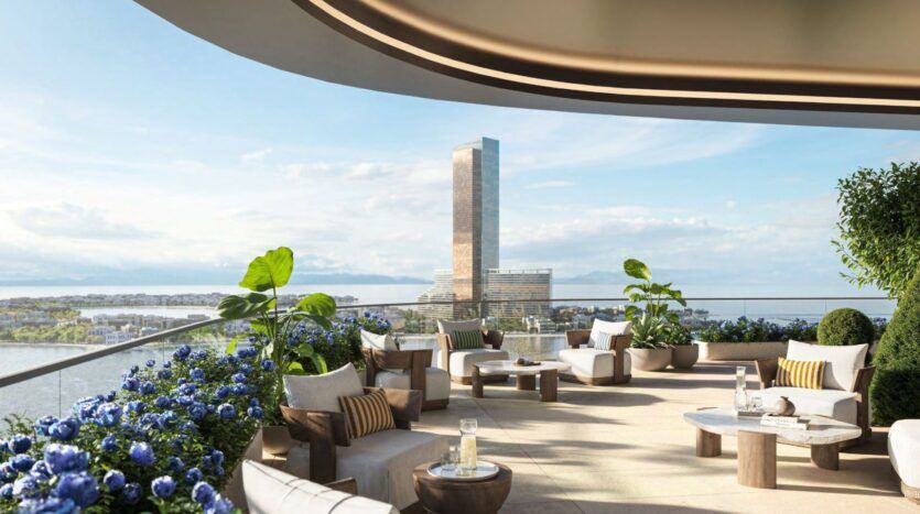 The Château's modern terrace, with its elegant seating, lush plants, and blue flowers, overlooks a river and a large skyscraper reflecting under a partly cloudy sky. The scene is bright, spacious, and welcoming.