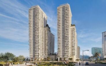 Deux tours résidentielles modernes avec balcons s'élèvent à Muheira, entourées de verdure, de gens qui marchent et de bâtiments urbains en arrière-plan, sous un ciel bleu clair.