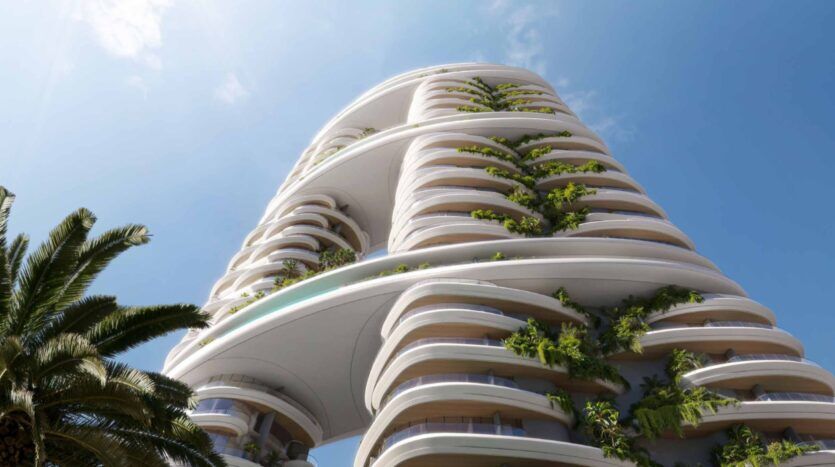 Amali Residences est un immeuble moderne de grande hauteur avec des balcons incurvés, chacun bordé de plantes vertes luxuriantes, se détachant sur un ciel bleu. Un palmier est visible au premier plan sur le côté gauche.