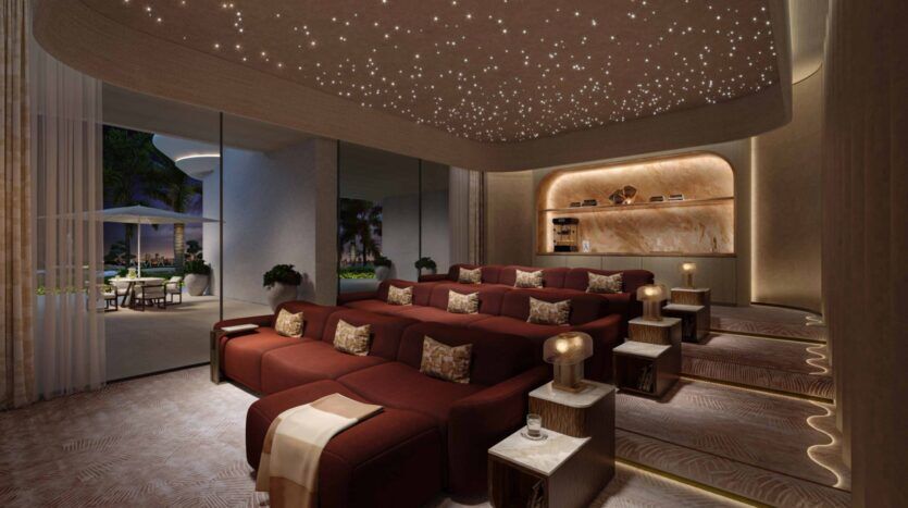 La luxueuse salle de cinéma des résidences Amali comprend des sièges rouges en gradins, de petites tables d'appoint avec des lampes et un plafond étoilé. Des fenêtres allant du sol au plafond révèlent un patio orné de palmiers et de meubles d'extérieur élégants.