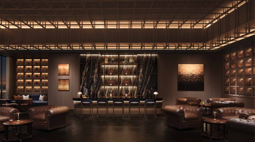 Un bar-salon sophistiqué dans les résidences Amali, avec des canapés et des fauteuils en cuir brun, un bar en marbre avec des tabourets, un éclairage d'ambiance chaleureux, des œuvres d'art murales abstraites et un mur d'étagères où sont exposés des bouteilles et des objets décoratifs.