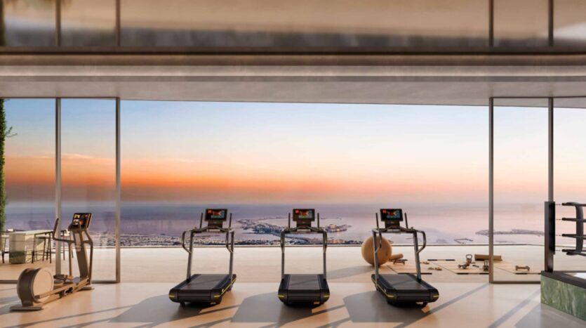Trois tapis de course font face à de grandes fenêtres allant du sol au plafond et offrant une vue panoramique sur la mer et la ville au coucher du soleil dans une salle de sport moderne des résidences Amali, dotée d'équipements élégants et d'une atmosphère sereine et luxueuse.