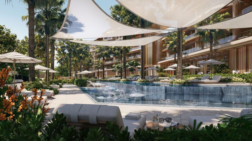 La luxueuse piscine extérieure de Four Seasons Private Residences, entourée de palmiers, de chaises longues, de parasols et de bâtiments modernes, avec des voiles d'ombrage au-dessus de la tête et de la verdure luxuriante, crée une atmosphère de station balnéaire relaxante.