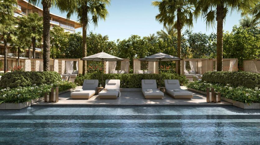 Scène de luxe au bord de la piscine de Four Seasons Private Residences, avec des chaises longues rembourrées, des parasols et des cabanas entourés de verdure luxuriante et de palmiers, par une journée ensoleillée avec un ciel bleu clair.