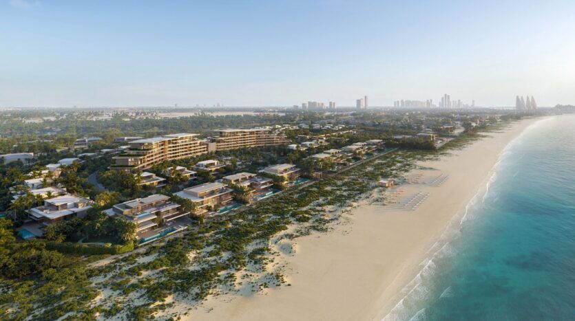 Vue aérienne de la station balnéaire Four Seasons Private Residences, dont les bâtiments modernes sont entourés de verdure, d'une plage de sable et d'un océan bleu. Des chaises longues et des parasols bordent le rivage ; la silhouette d'une ville est visible à l'arrière-plan.