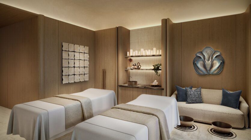 La salle de spa sereine de Four Seasons Private Residences comprend deux tables de massage, du linge beige, un canapé moderne avec des oreillers bleus, une étagère avec des bougies et des objets de décoration, ainsi qu'un éclairage doux et chaleureux pour créer une atmosphère vraiment relaxante.