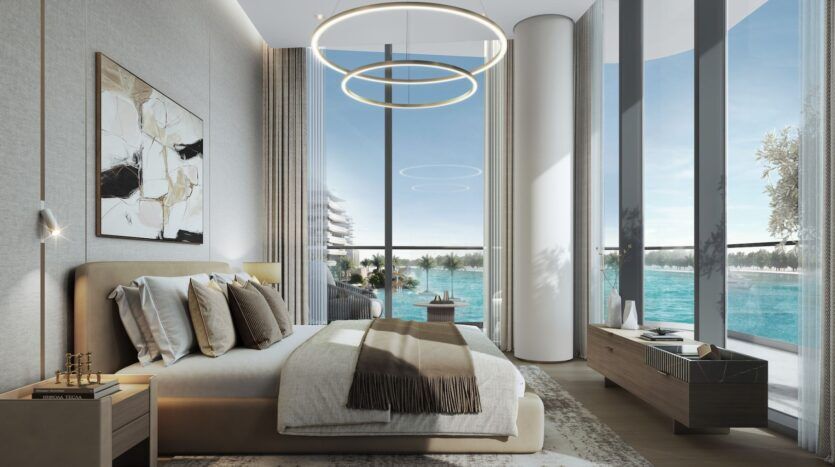 Chambre moderne à River Cove Residences avec des fenêtres allant du sol au plafond et donnant sur un front de mer bleu, un lit beige avec plusieurs oreillers, des œuvres d'art murales abstraites, des plafonniers circulaires et des meubles en bois minimalistes.