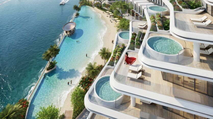 Vue aérienne de River Cove Residences, un complexe de luxe en bord de mer avec des piscines privées sur les balcons, une plage de sable, des palmiers et des personnes nageant dans un lagon bleu au bord de la mer. Un yacht est visible dans l'eau.