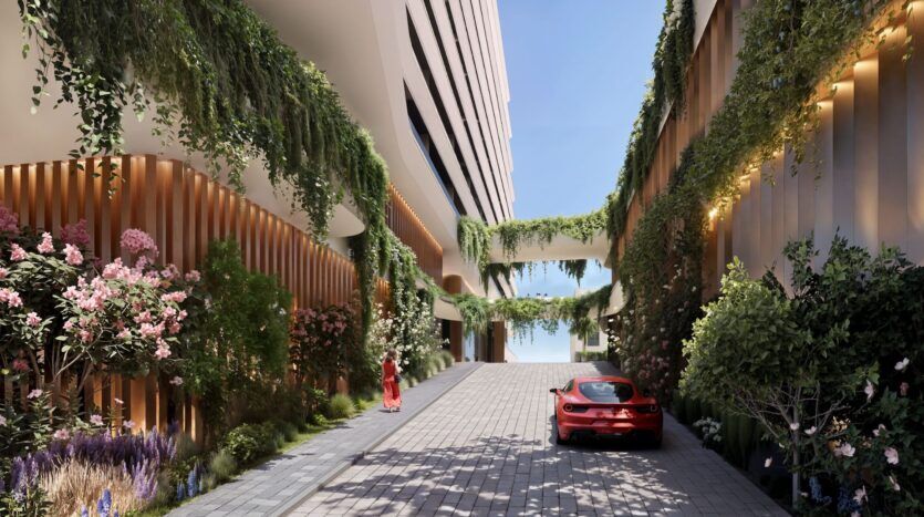 Une cour moderne des résidences River Cove, avec de grands bâtiments ornés de verdure et de fleurs luxuriantes. Une voiture de sport rouge est garée sur le chemin de pierre, tandis qu'une personne en rouge marche vers le ciel bleu ouvert au-delà de la cour.