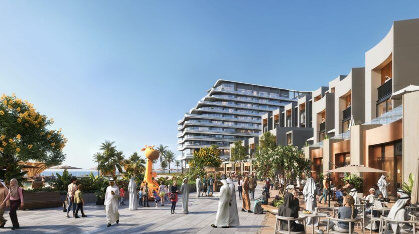 A lively outdoor promenade at The Orchard by Sobha, with people strolling and enjoying a bite to eat at cafés, palm trees, modern buildings, and a large inflatable giraffe under a clear blue sky.