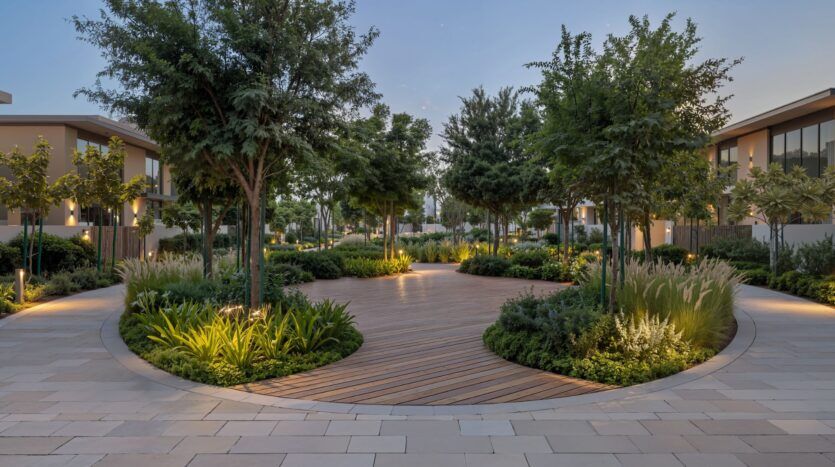 The landscaped courtyard at The Orchard by Sobha features a circular wooden walkway, surrounded by green trees, shrubs, and modern buildings on either side, all softly illuminated by outdoor lighting at dusk.