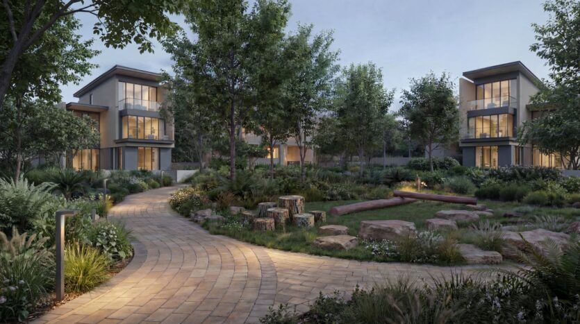 A landscaped garden with a winding stone path, surrounded by trees, shrubs, and modern two-story buildings at dusk at The Orchard by Sobha. Logs and stones create a cozy spot to sit in this peaceful setting.
