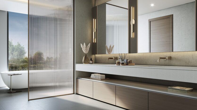 A modern bathroom at The Orchard by Sobha featuring a double-sink vanity, large mirrors, warm lighting, built-in cabinets, and a freestanding bathtub next to a large window. The neutral tones and minimalist decor create a clean and serene atmosphere.
