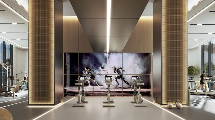 The Orchard by Sobha’s modern fitness center features tall columns, exercise bikes facing a large screen showing boxing matches, and a variety of fitness equipment along the walls. The interior is bright, spacious, and well-lit, with a contemporary design.