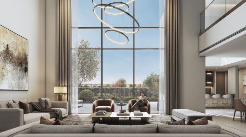 A spacious, modern living room at The Orchard by Sobha, featuring floor-to-ceiling windows, a contemporary circular chandelier, neutral-toned sofas, armchairs, a large coffee table, wall art, and a view of the garden. A person is sitting peacefully by the window, reading.