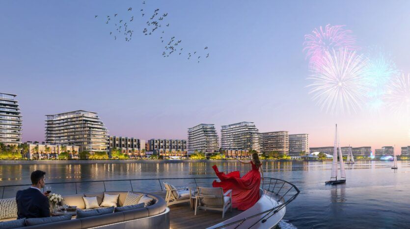 A woman wearing a red dress is sitting on a yacht at sunset, facing a waterfront cityscape reminiscent of The Orchard by Sobha. Fireworks light up the sky, birds fly overhead, a sailboat bobs nearby, and a man is sitting at a table on the yacht.
