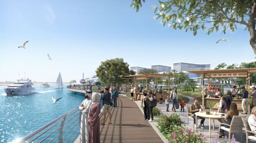 Les gens se promènent le long de The Terraces by Sobha, une promenade moderne au bord de l'eau, bordée de cafés, d'arbres et de sièges en plein air. Les bateaux glissent sur l'eau bleue tandis que les mouettes s'envolent, créant une scène animée sous un ciel clair et ensoleillé.