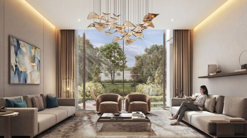 Un salon moderne aux tons neutres à The Terraces by Sobha est doté de grandes fenêtres donnant sur un jardin. Des meubles élégants, des œuvres d'art abstraites et un lustre doré décorent l'espace. Une femme est assise sur un canapé et lit un livre.