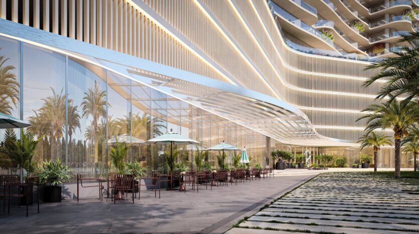 An outdoor terrace with tables, chairs, and umbrellas next to the modern glass structure of the Wow Tower, featuring palm trees, potted plants, and undulating architectural lines under a clear sky.