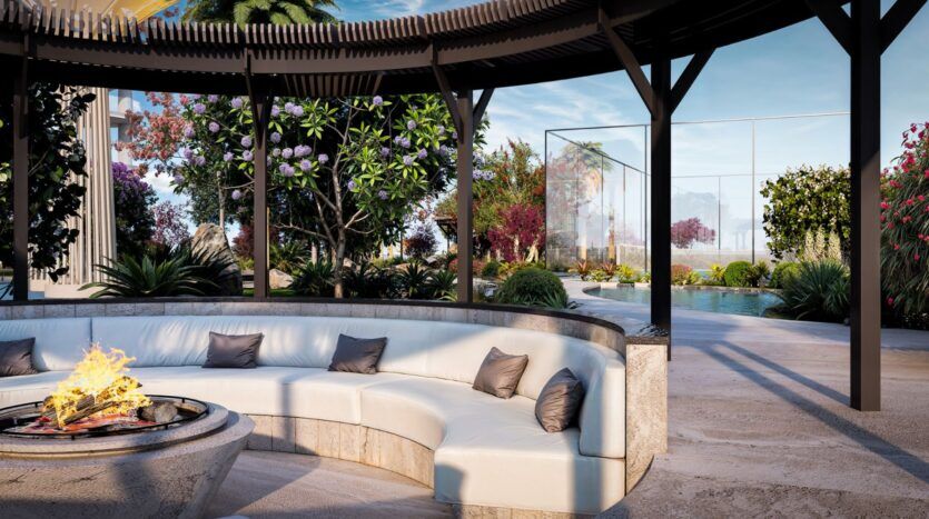 The modern outdoor living area at Wow Tower features a curved white sofa with gray cushions under a pergola, a fireplace in front, lush greenery, blooming flowers, and a swimming pool with a transparent glass fence in the background.