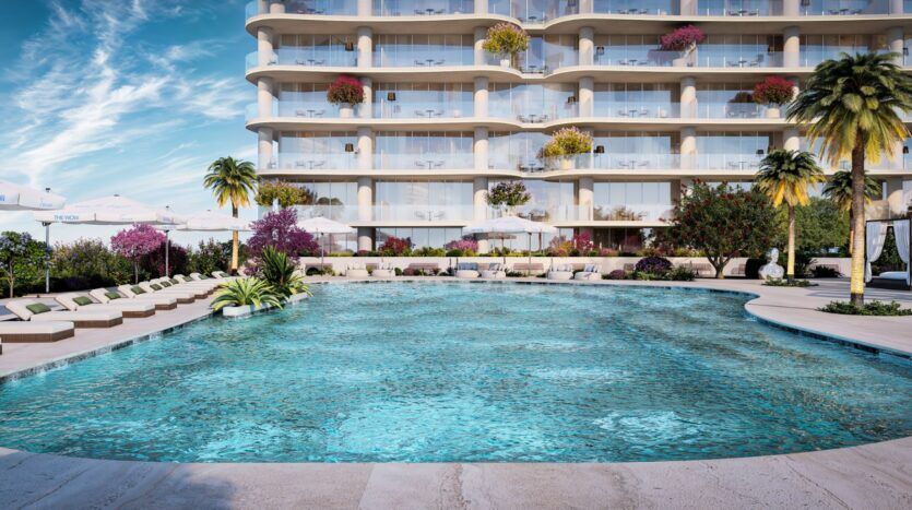 A luxurious hotel pool with crystal-clear blue water is nestled beneath the Wow Tower, surrounded by lounge chairs, umbrellas, palm trees, and colorful flowers. Balconies adorned with plants overlook the pool beneath a blue sky dotted with clouds.