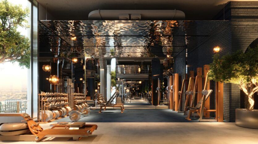 The modern, spacious gym at the Wow Tower is equipped with wooden fitness equipment, free weights, benches, and floor-to-ceiling windows that flood the sleek interior with natural light. Green plants add a touch of freshness to this elegant space.