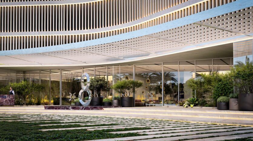 The modern entrance to The Wow Tower features large floor-to-ceiling windows, a curved metal canopy, decorative lighting, potted plants, a contemporary sculpture, and meticulously landscaped grounds in the foreground.