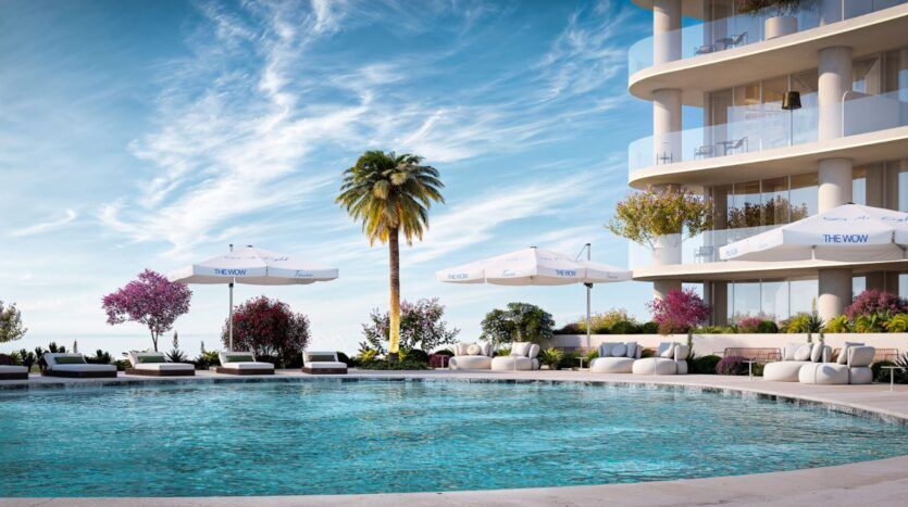 The Wow Tower, a modern, curved building with balconies, overlooks a circular outdoor pool surrounded by lounge chairs, white umbrellas, palm trees, and brightly blooming flowers under a brilliant blue sky.