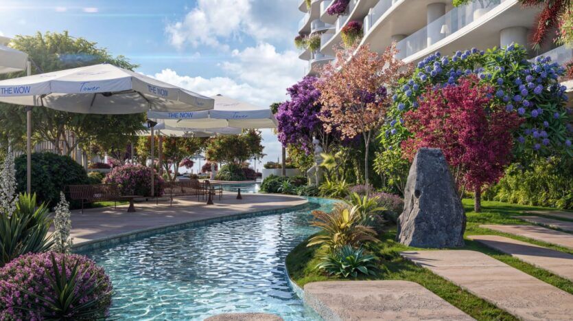 The landscaped courtyard of the Wow Tower features a clear stream surrounded by lush greenery, colorful flowers, and trees. Benches and shaded seating areas with umbrellas line the path alongside the modern, curved building.