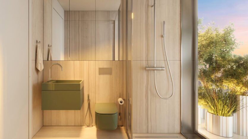 A modern bathroom with light wood walls inside the Wow Tower, featuring a green floating sink and matching toilet, a walk-in glass shower, and large plants next to a window that lets in natural sunlight.
