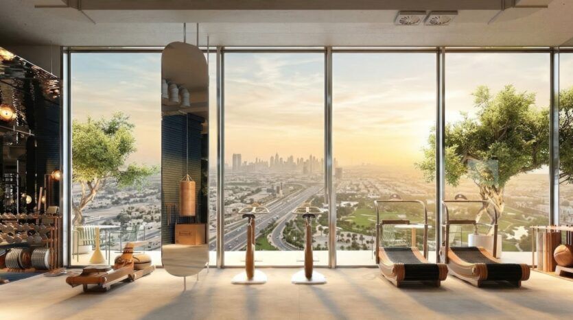 A modern, bright fitness center at the Wow Tower featuring exercise equipment, large floor-to-ceiling windows, potted trees, wood accents, and a panoramic view of the city at sunrise or sunset.