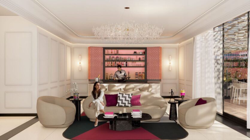 Un luxueux salon de la résidence Fauchon présente des murs couleur crème et un mobilier moderne. Une femme est assise sur un canapé beige, un verre à la main, tandis qu'un barman se tient derrière un bar rétroéclairé. Un lustre est suspendu au-dessus du bar, tandis que la lumière du soleil pénètre par la droite.