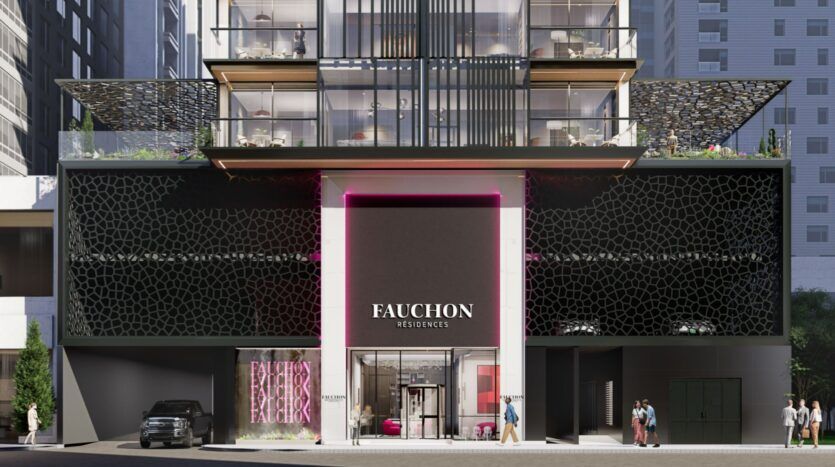 Extérieur d'un bâtiment moderne avec une façade géométrique noire, une grande enseigne indiquant "Résidence Fauchon", une entrée vitrée et des espaces commerciaux au niveau de la rue. Des personnes et une voiture garée sont visibles devant la résidence Fauchon.