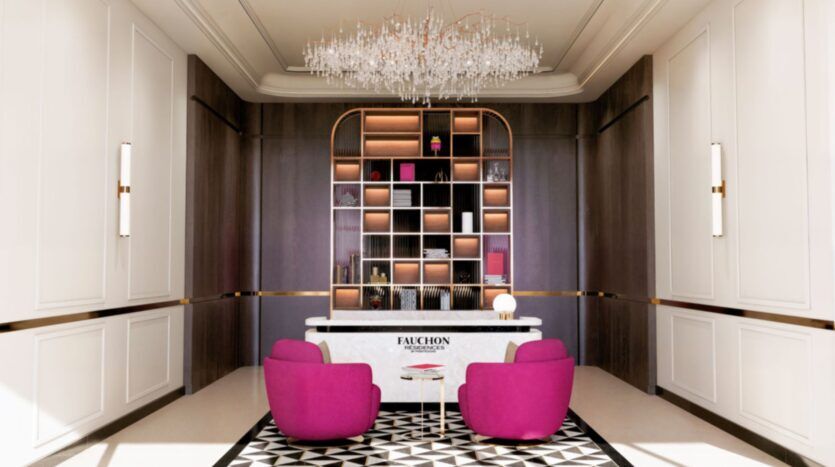 L'espace de réception moderne de la résidence Fauchon comprend deux chaises rose vif, un tapis géométrique noir et blanc, un bureau en marbre portant l'inscription "Fauchon", une haute étagère rétroéclairée et un lustre en cristal au dessus de la tête.