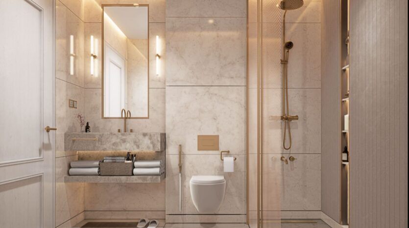 La salle de bains moderne de la résidence Fauchon présente des murs en marbre, un grand miroir au-dessus d'un lavabo flottant, des toilettes murales, une douche en verre avec des accessoires en or, une étagère à serviettes et un éclairage d'ambiance doux.