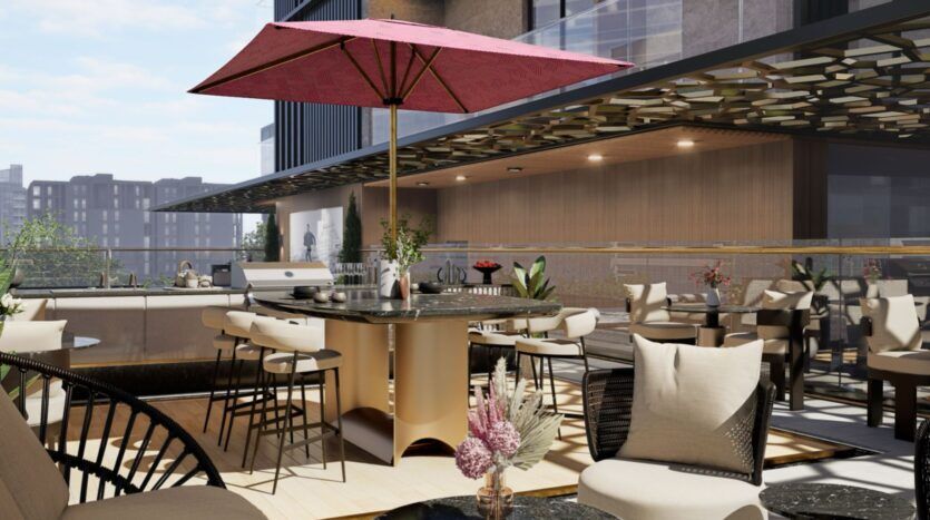 Un patio extérieur moderne de la résidence Fauchon avec des chaises et des tables beiges, un grand barbecue rond et un parasol rouge. Les bâtiments de la ville sont visibles à l'arrière-plan et des plantes en pot ajoutent de la verdure à ce cadre élégant.