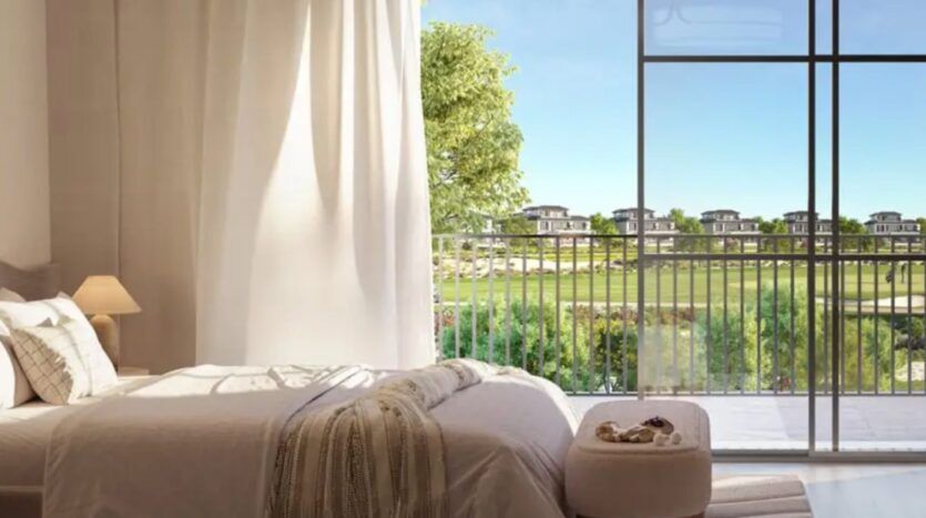Une chambre lumineuse avec un grand lit, des draps doux et une lampe de chevet, dotée de fenêtres allant du sol au plafond et donnant sur le balcon et la verdure de Hudayriyat Golf Estates, avec des maisons modernes au loin sous un ciel bleu clair.