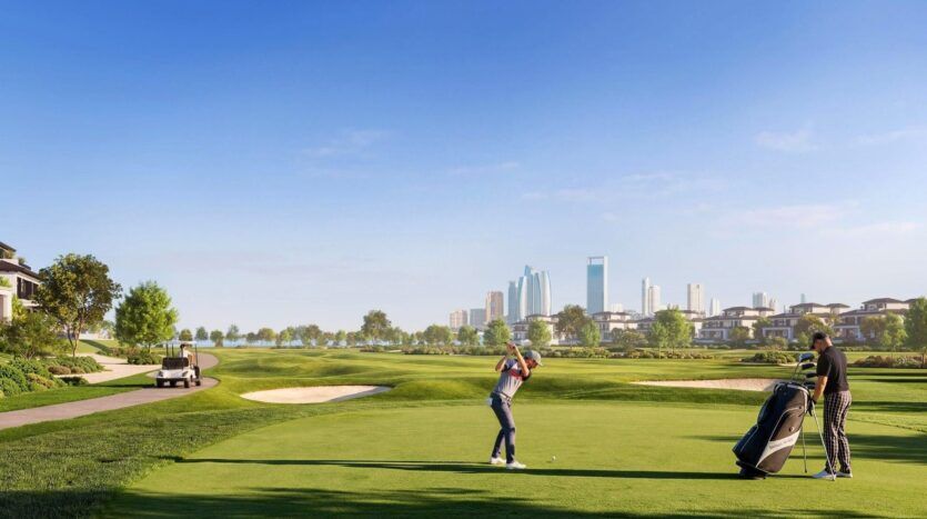 Deux personnes jouent au golf à Hudayriyat Golf Estates sur un parcours verdoyant avec des bunkers de sable, une voiturette de golf à proximité et des gratte-ciel modernes en arrière-plan sous un ciel bleu clair.