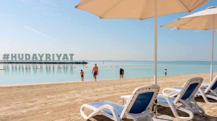 Plusieurs chaises longues vides sous des parasols bordent la plage de sable de Hudayriyat Golf Estates, avec des gens qui se promènent vers une eau calme et bleue. De grandes lettres épellent "#HUDAYRIYAT" sur une plateforme dans l'eau sous un ciel clair.
