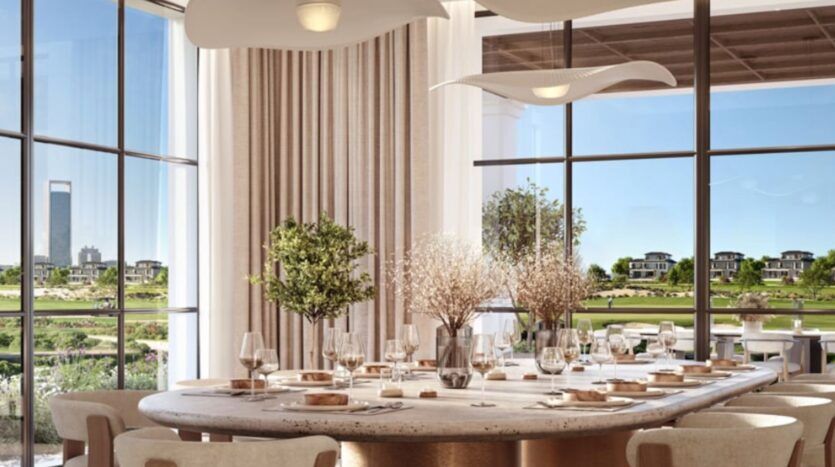 Salle à manger élégante avec une grande table ronde dressée pour un repas, décorée de verrerie, d'assiettes et de centres de table floraux. Les grandes fenêtres offrent une vue sur les pelouses vertes et les maisons modernes de Hudayriyat Golf Estates.