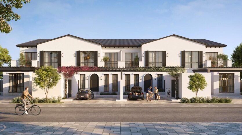 Complexe moderne de maisons de ville blanches de deux étages avec de grandes fenêtres, des balcons et des arches à Hudayriyat Golf Estates. Deux voitures sont garées devant, des gens se tiennent près de l'entrée et une personne fait du vélo sur les trottoirs bordés d'arbres.