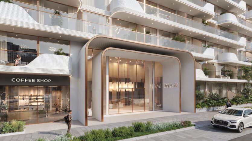 L'extérieur de l'immeuble est moderne avec des balcons incurvés, des garde-corps en verre et des jardinières paysagées. L'entrée étiquetée "MARI IMAN" vous accueille à Oxford Cove by Iman. Un café avec des sièges est visible sur la gauche, à côté de piétons et d'une voiture à proximité.