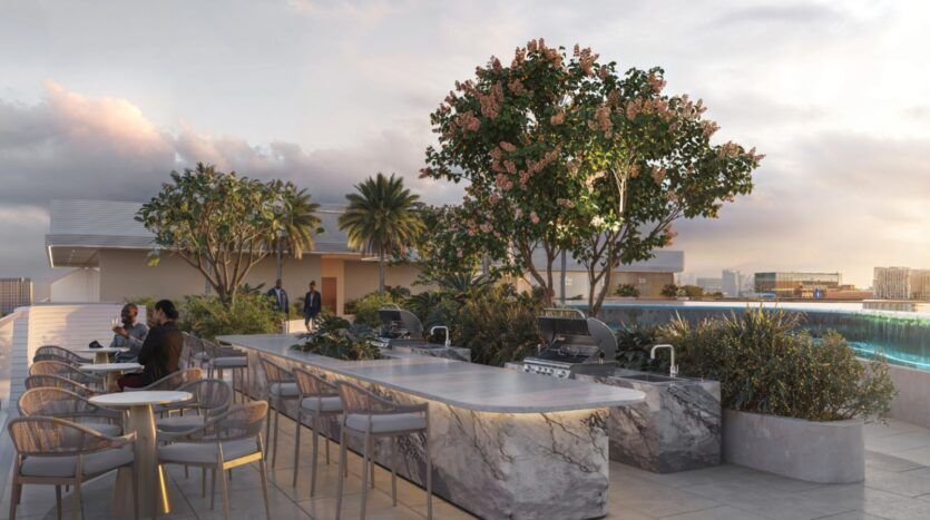 Le toit-terrasse moderne de l'Oxford Cove by Iman est doté de comptoirs en marbre et de grills intégrés, entourés de tables et de chaises, d'une végétation luxuriante et d'une vue sur la ville sous un ciel partiellement nuageux au coucher du soleil.
