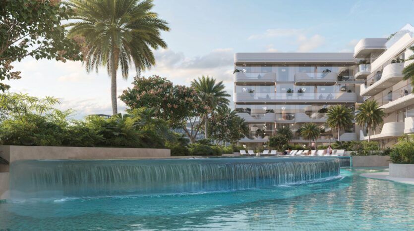 L'immeuble d'appartements de luxe moderne Oxford Cove by Iman est doté de balcons en verre donnant sur une piscine sereine avec une cascade, entourée d'une végétation luxuriante, de palmiers et de chaises longues sous un ciel bleu.