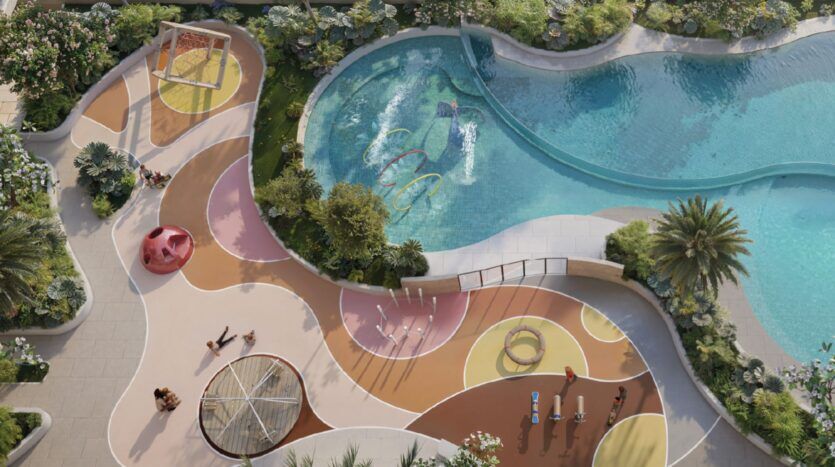 Vue aérienne d'une aire de jeux colorée à côté d'une piscine incurvée à Oxford Cove by Iman, avec des toboggans, des structures d'escalade, des motifs circulaires, et des enfants jouant tandis que les adultes sont assis à proximité au milieu d'une végétation luxuriante.