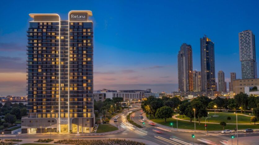 Paysage urbain en soirée montrant un grand hôtel bien éclairé portant le nom de "Rotana Residences", avec des voitures créant des traînées lumineuses sur une route sinueuse, entouré d'arbres, d'autres gratte-ciel et d'un ciel bleu au crépuscule.