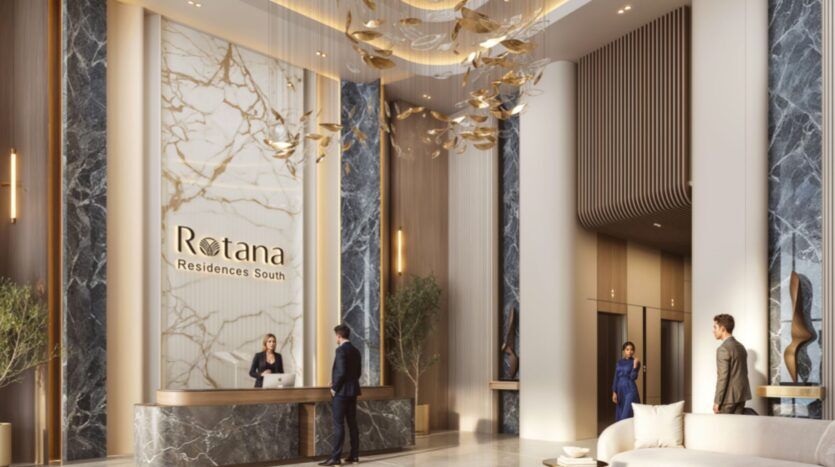 Le hall d'entrée moderne et élégant de l'hôtel Rotana Residences est orné de murs en marbre, de touches dorées, d'un lustre sculptural et de la signalétique "Rotana Residences South". Un réceptionniste assiste un client tandis que d'autres passent devant des ascenseurs et des sièges élégants.