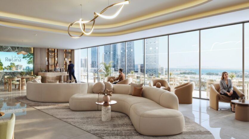 Le salon moderne de l'hôtel Rotana Residences est composé de canapés beiges incurvés, de tables rondes, de grandes fenêtres offrant une vue sur les gratte-ciel de la ville et sur l'eau, d'un bar et de personnes se relaxant dans un espace lumineux et aéré.