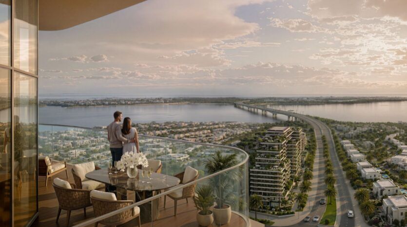 Un couple se tient sur le balcon d'une tour moderne de Rotana Residences qui surplombe une rivière, un long pont et le paysage urbain au coucher du soleil. Le balcon comprend une table à manger, des chaises, des plantes en pot et une vue étendue sur l'eau et les bâtiments.